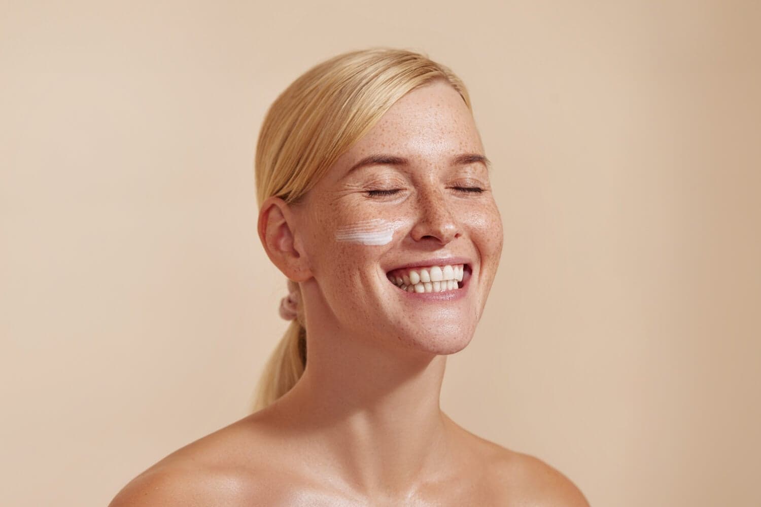 Smiling blonde woman with freckles and closed eyes, a stripe of white cream on her cheek, against a beige backdrop.