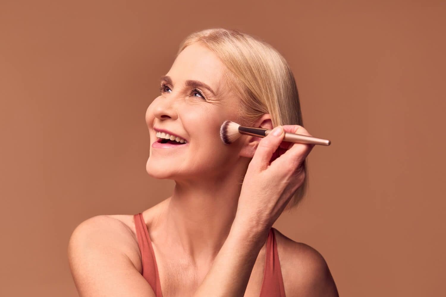 Smiling blonde woman in a rust tank top sweeping a gold-handled powder brush along her cheekbone against a warm brown backdrop.