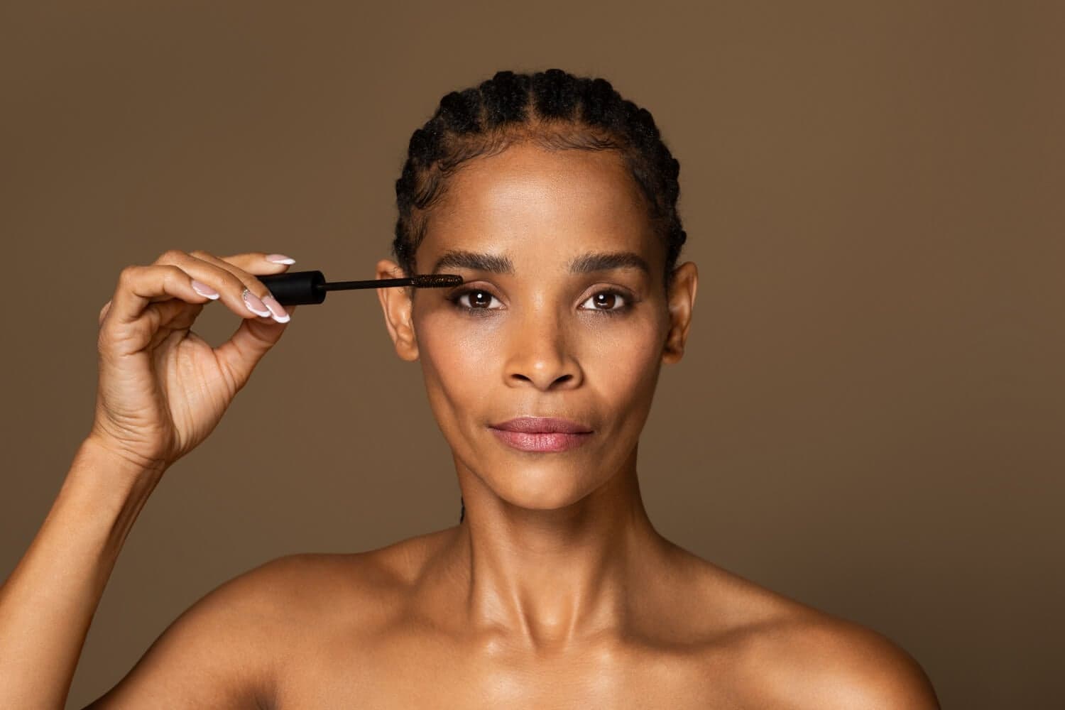 Woman with cornrow braids holding an unbranded black mascara wand with a brown-coated brush to her lashes. Woman with cornrow braids holding an unbranded black mascara wand with a brown-coated brush to her lashes.