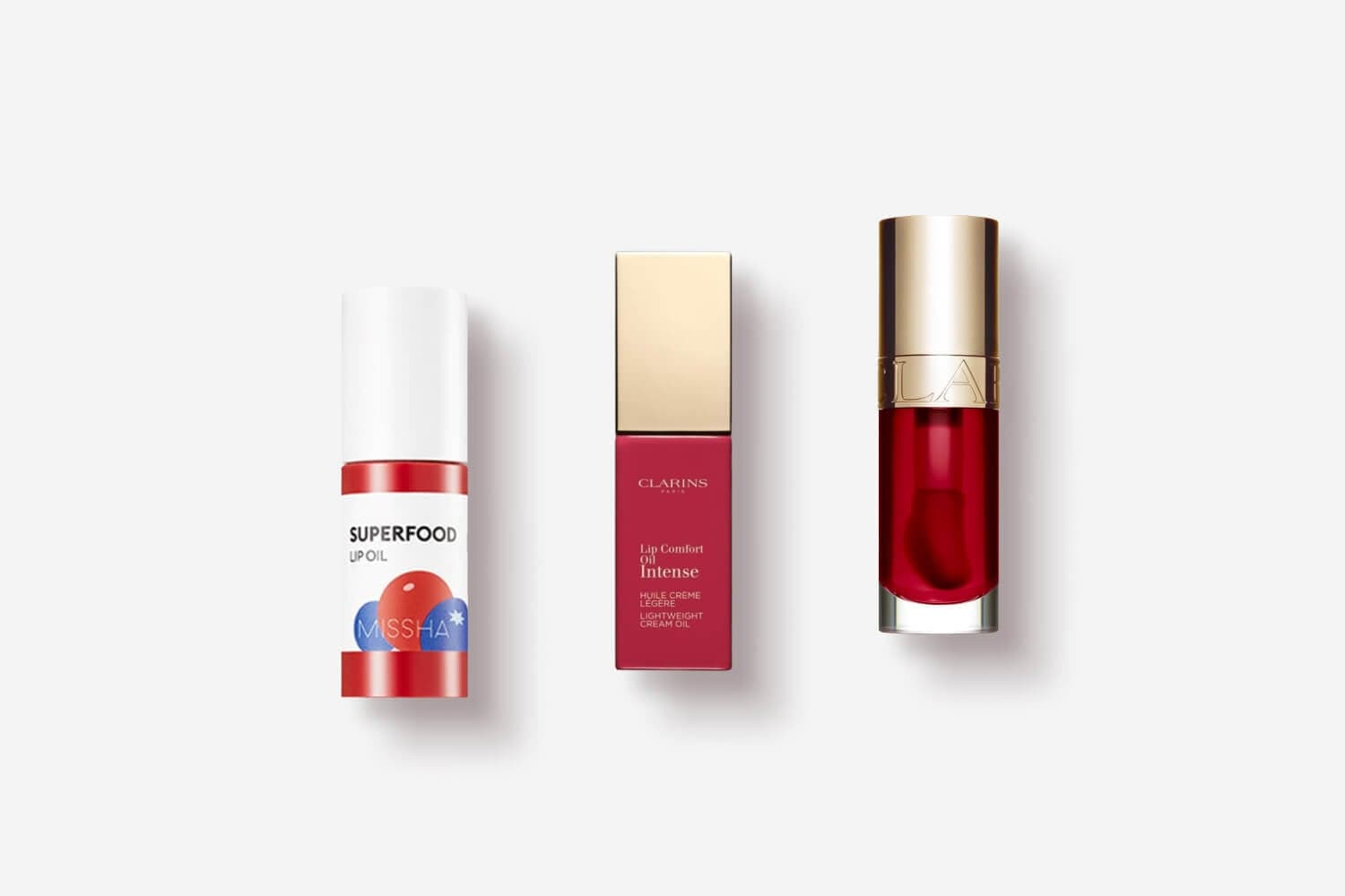 Missha Superfood Lip Oil beside Clarins Lip Comfort Oil Intense in its box and a red gold-capped bottle. Missha Superfood Lip Oil beside Clarins Lip Comfort Oil Intense in its box and a red gold-capped bottle.