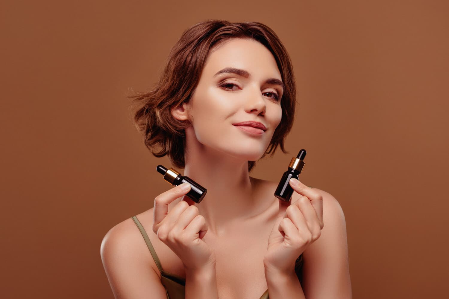 Smiling woman with a short brown bob holding two unlabelled amber glass serum dropper bottles against a warm brown backdrop. Smiling woman with a short brown bob holding two unlabelled amber glass serum dropper bottles against a warm brown backdrop.