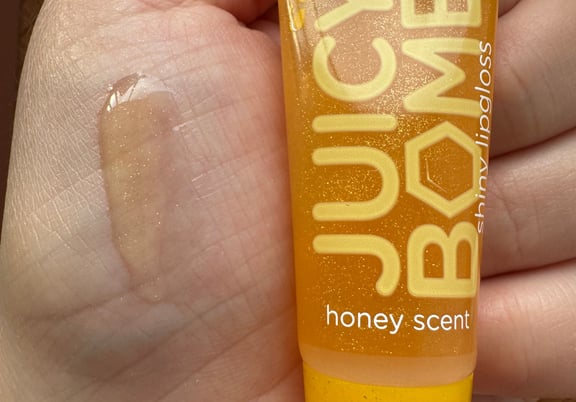 Yellow tube of Essence Juicy Bomb shiny lip gloss with honey scent, shown with clear gloss swatch on skin. review