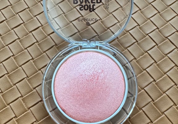 Round compact with clear lid containing shimmery pink baked blush on a textured tan surface. review
