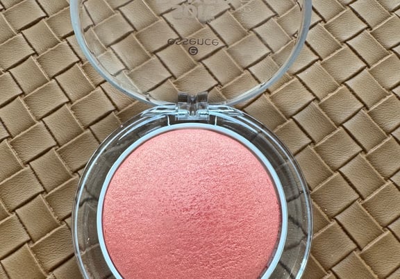 Round, pink baked blush in a clear plastic compact case, placed on a tan textured surface. review